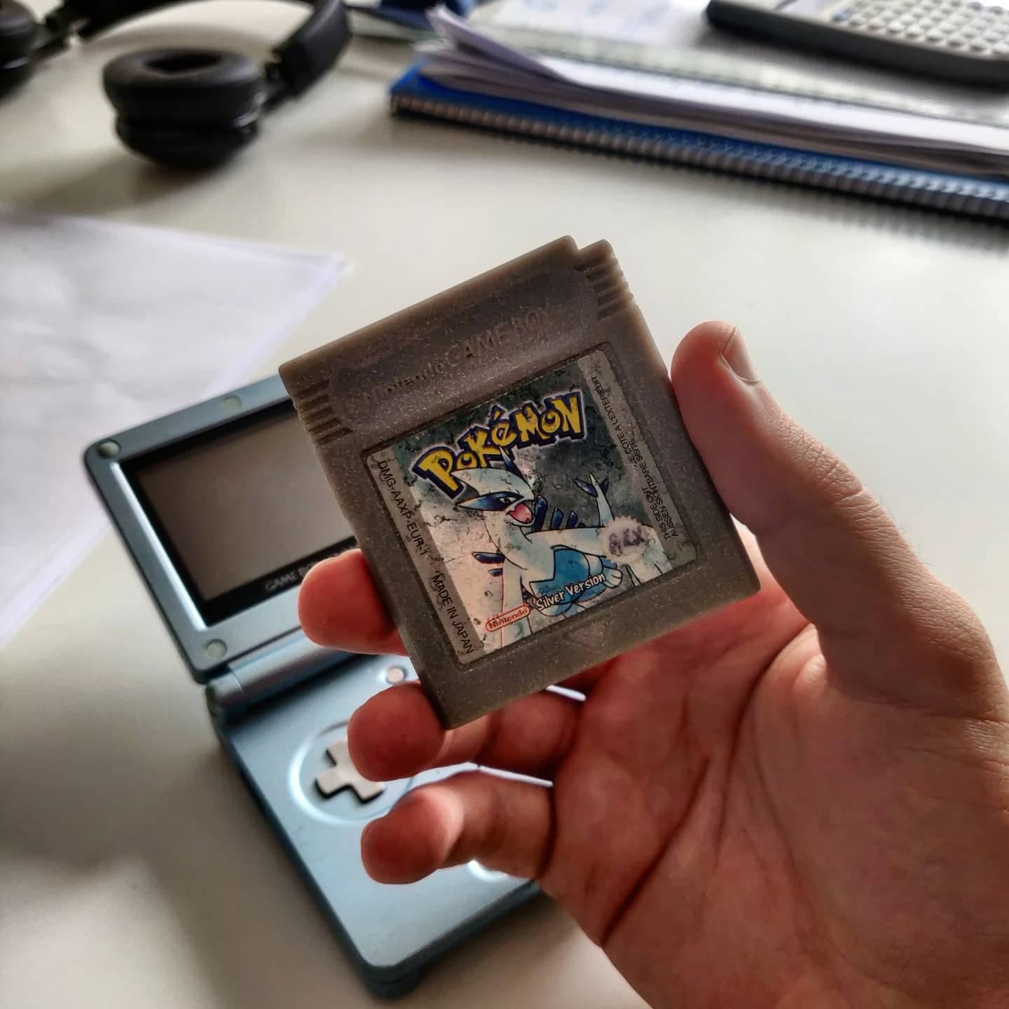 A hand holding a worn Pokémon cartridge above a Game Boy Advance SP
